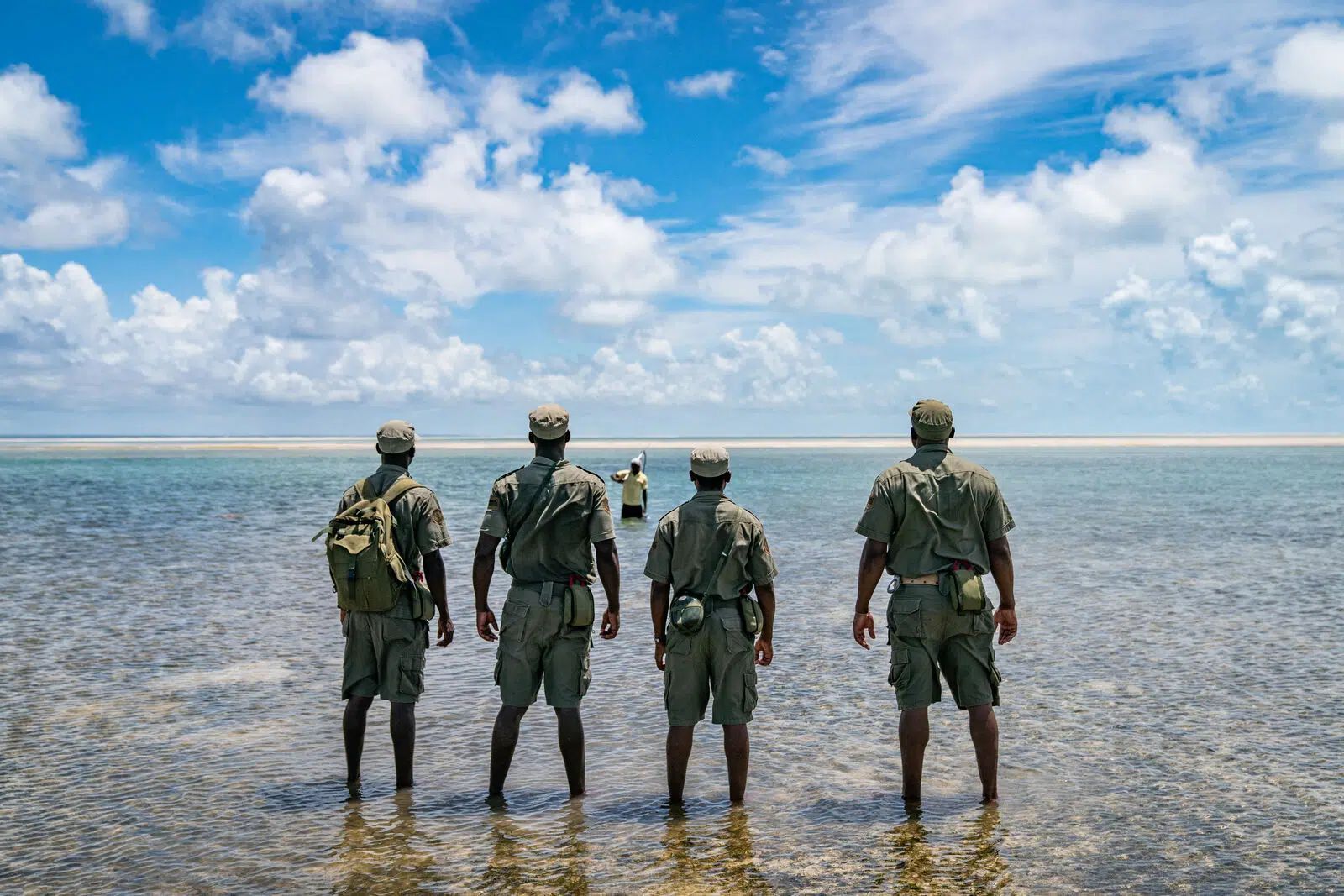 Four soldiers stand in shallow water facing the open sea under a cloudy blue sky.