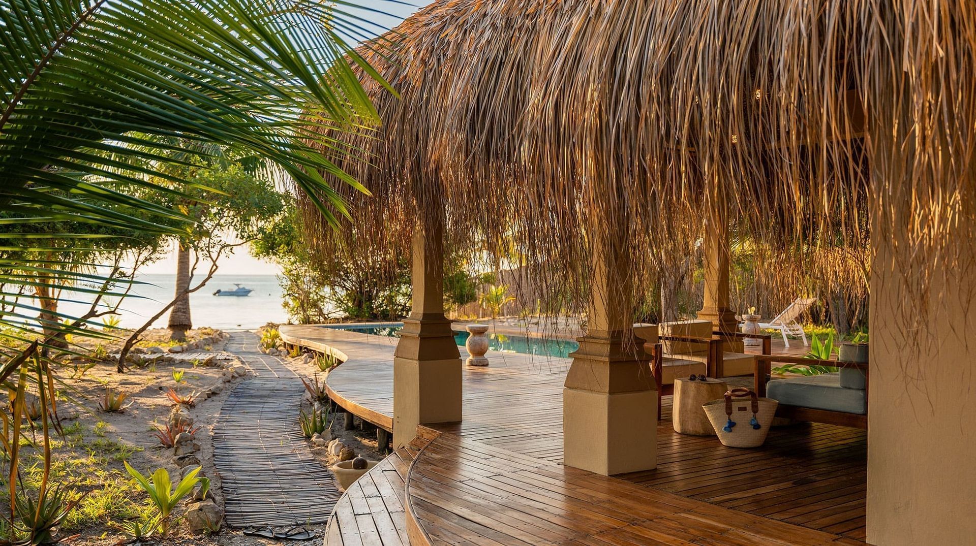 Beachside hut with thatched roof, palm fronds, and sunlit wooden deck beside the water