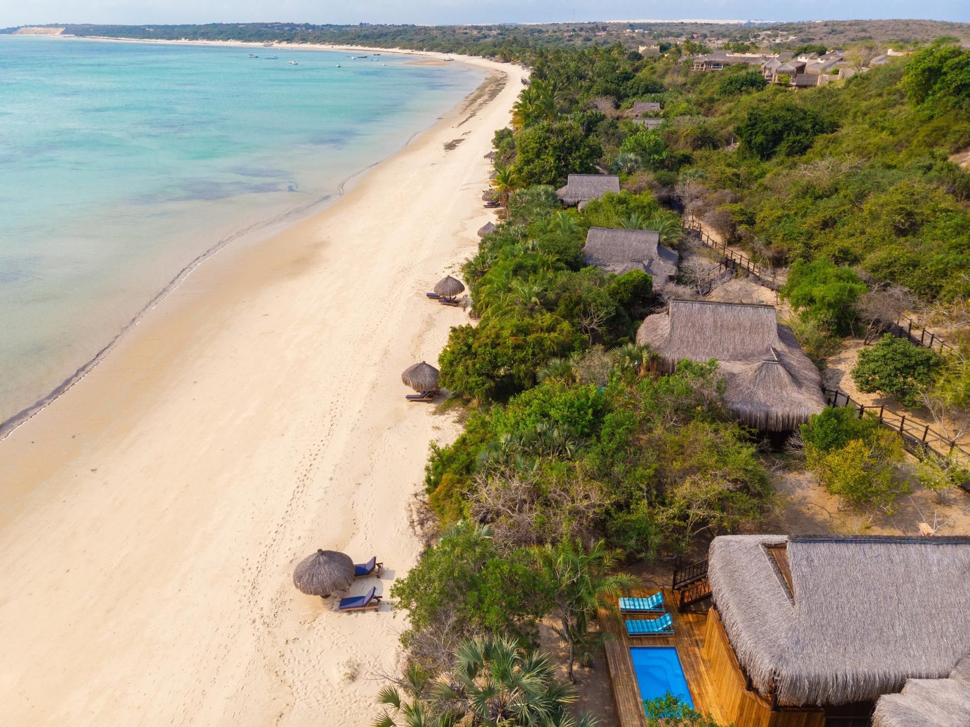 Aerial view of a tropical beach with turquoise water, white sand, and thatched huts beside dense greenery