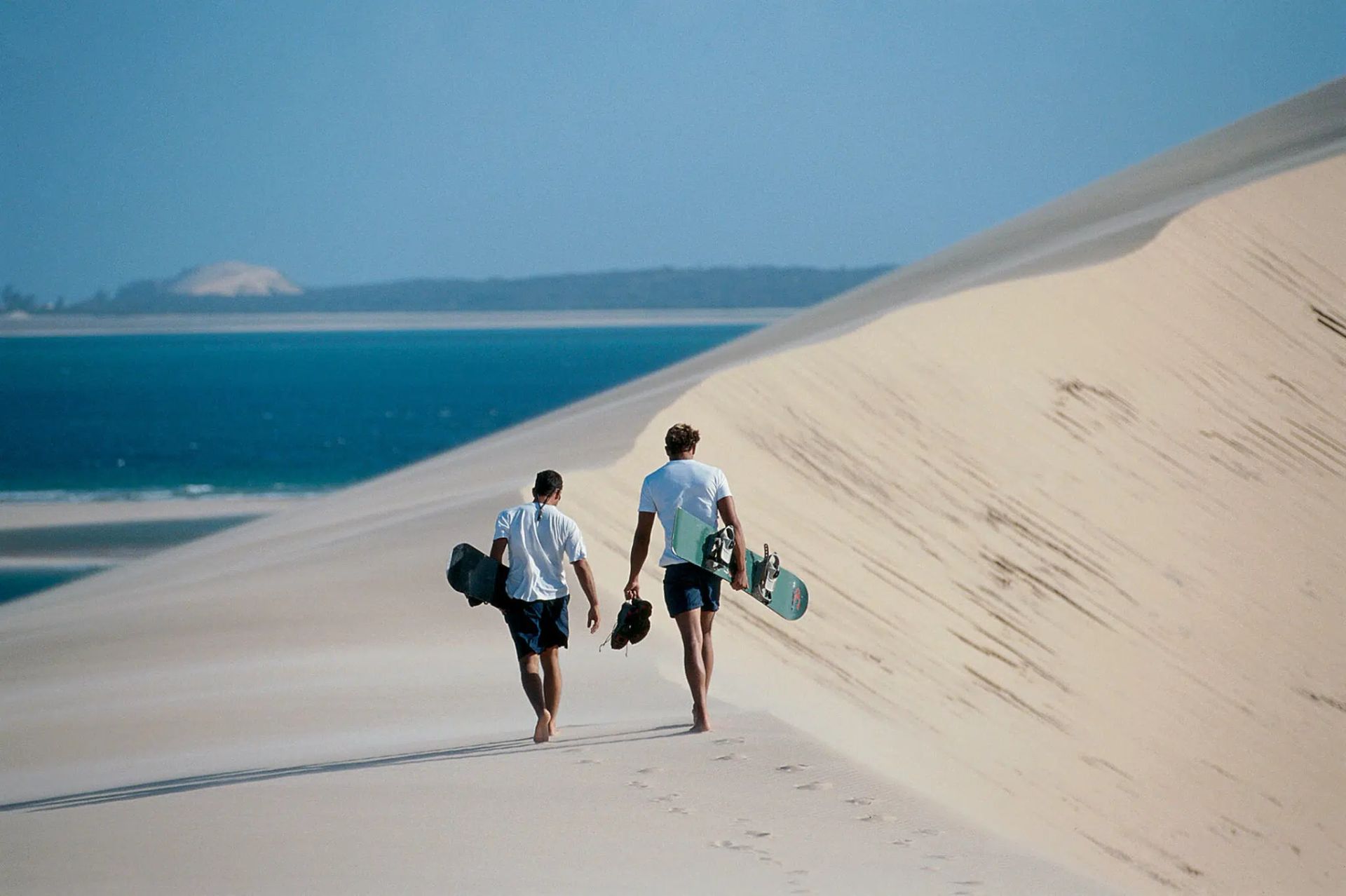 Two people walk along a sand dune beside a blue sea, carrying bags under a clear sky.