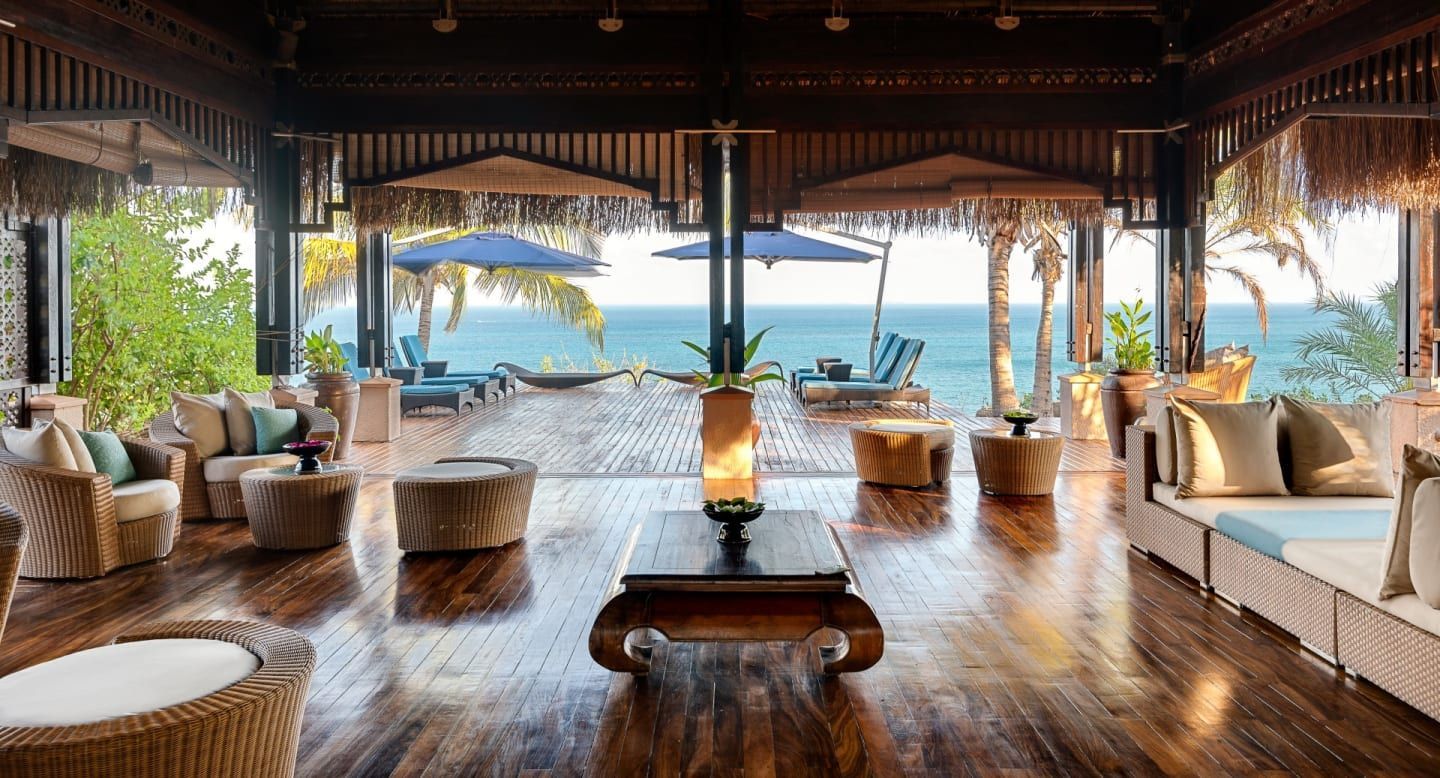 Open-air beachfront lounge with wicker seating, wooden deck, and ocean view through wide pillars