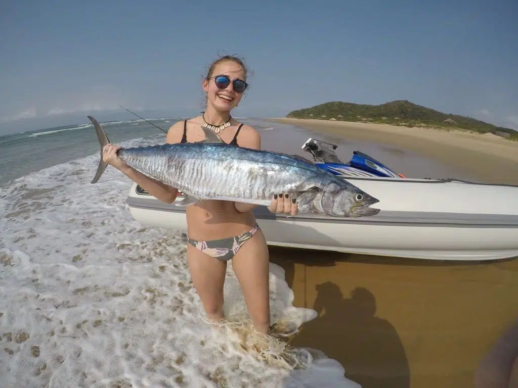Woman in sunglasses holding a large silver fish beside a boat on a sandy beach with waves breaking.