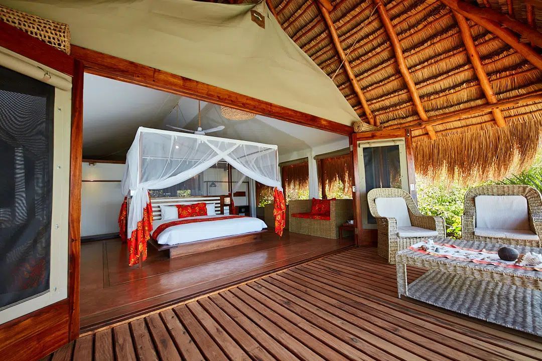 Open-air thatched bedroom with canopy bed, wooden deck, and lounge chairs overlooking greenery