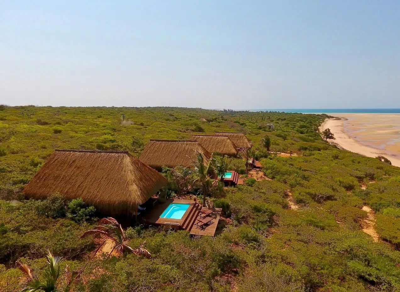 Thatched beach huts beside a turquoise pool and sandy shoreline in a green coastal landscape