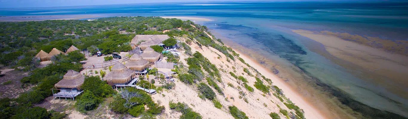 Aerial view of a sandy island with huts, green vegetation, and turquoise water along the shore.