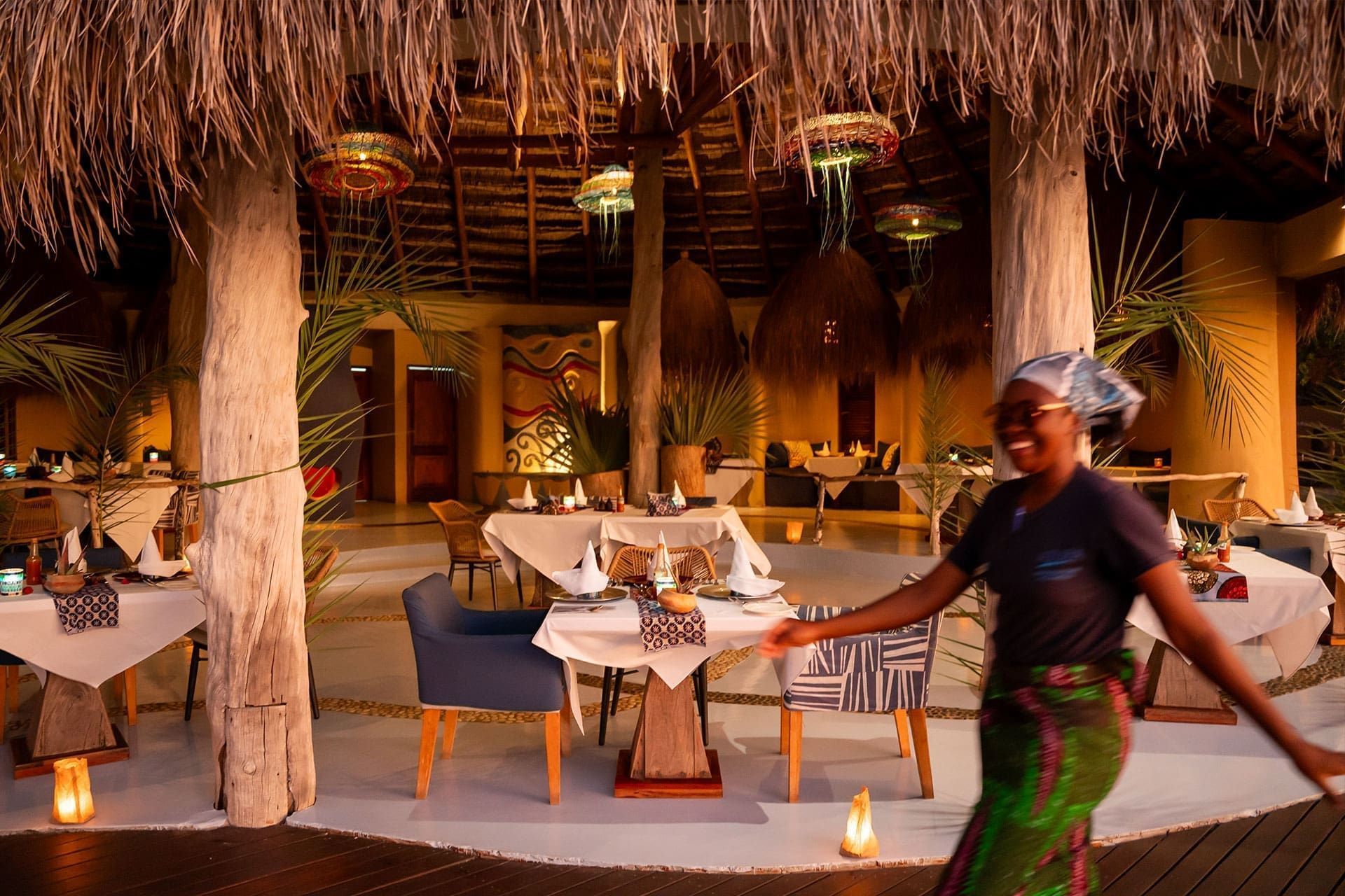 Tropical resort dining area with thatched roof, lit tables, and a server walking past