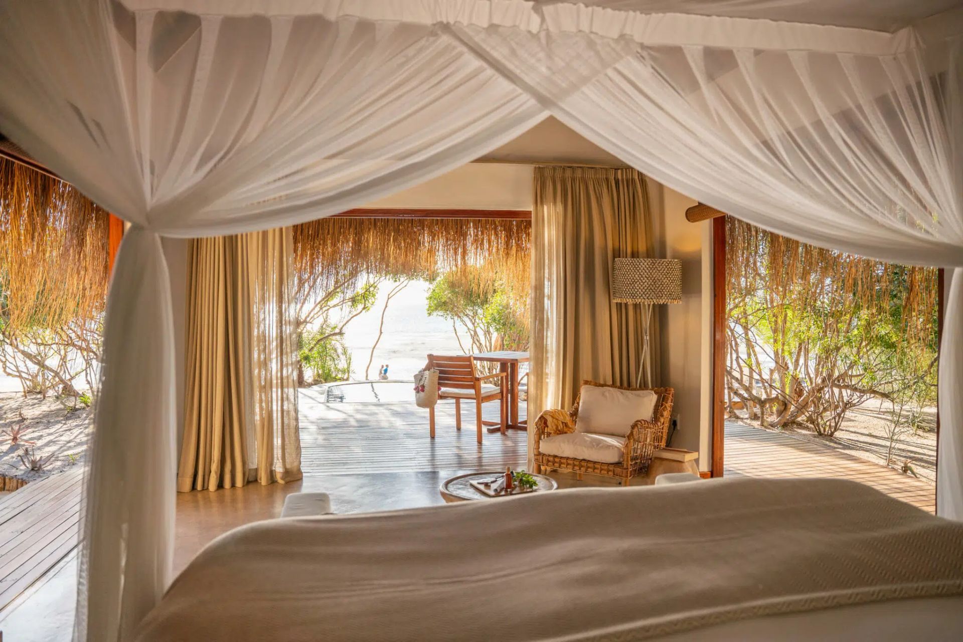 Canopy bed view of a beachside hut with white drapes, wicker chair, and ocean beyond