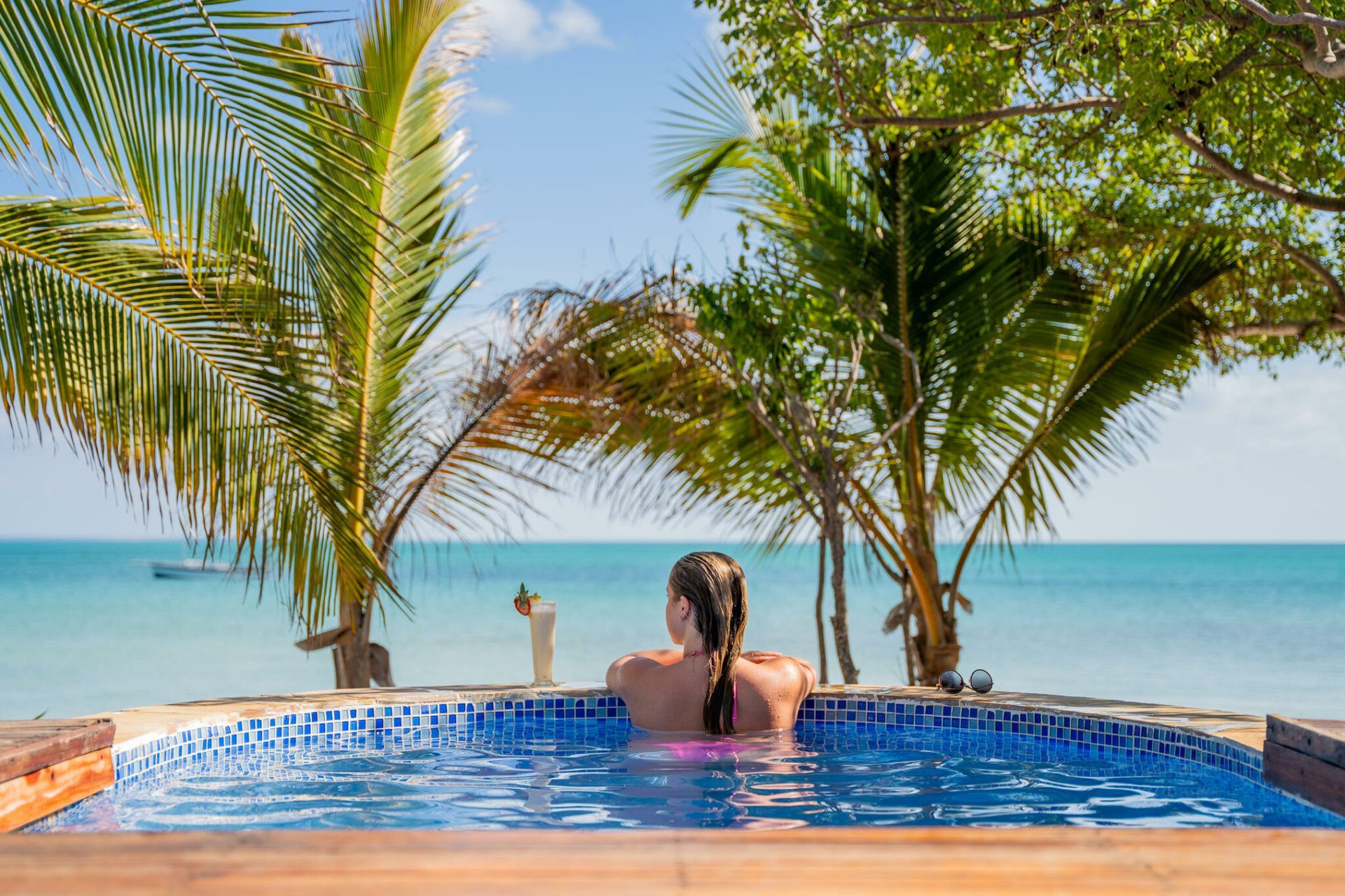 Person relaxing in a blue infinity pool overlooking the ocean, framed by palm trees and a sunny sky