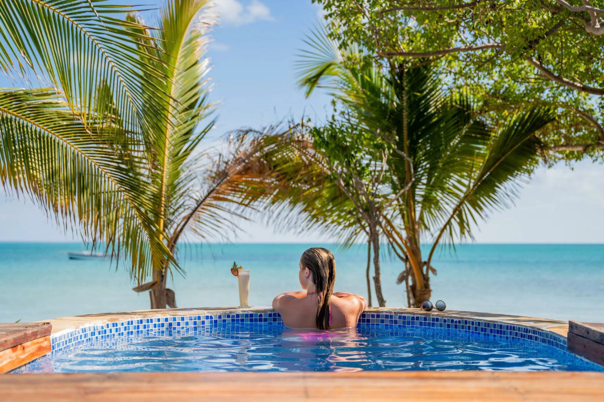 Person relaxing in a pool overlooking the ocean, framed by palm trees at sunset