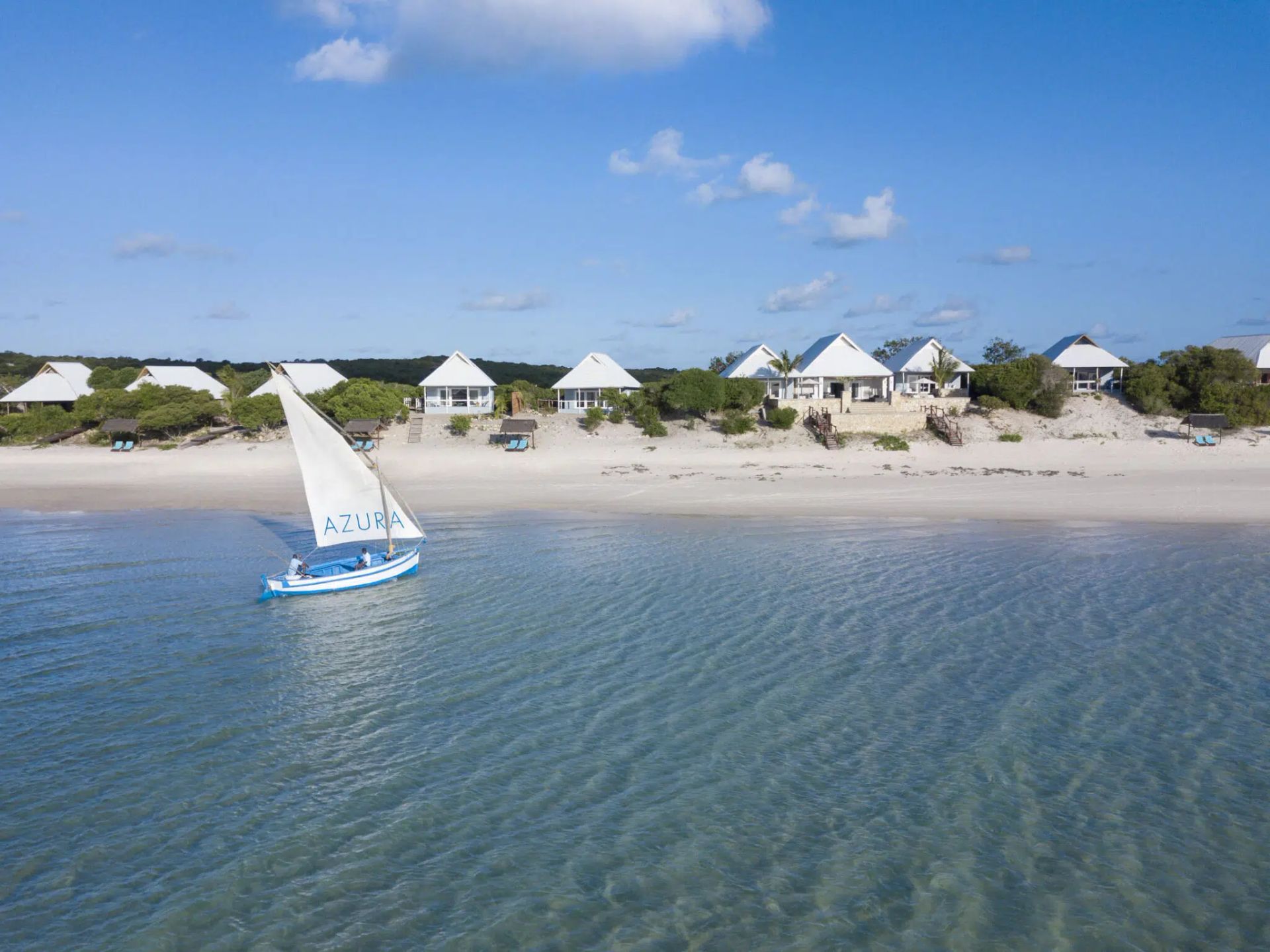 Blue sailboat on calm water near a sandy beach with white vacation homes under a clear sky