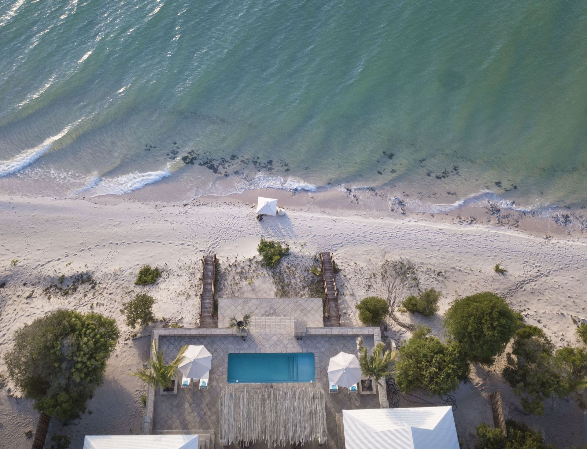 Aerial view of a beachfront pool, deck, and cabana beside green ocean water and sandy shore