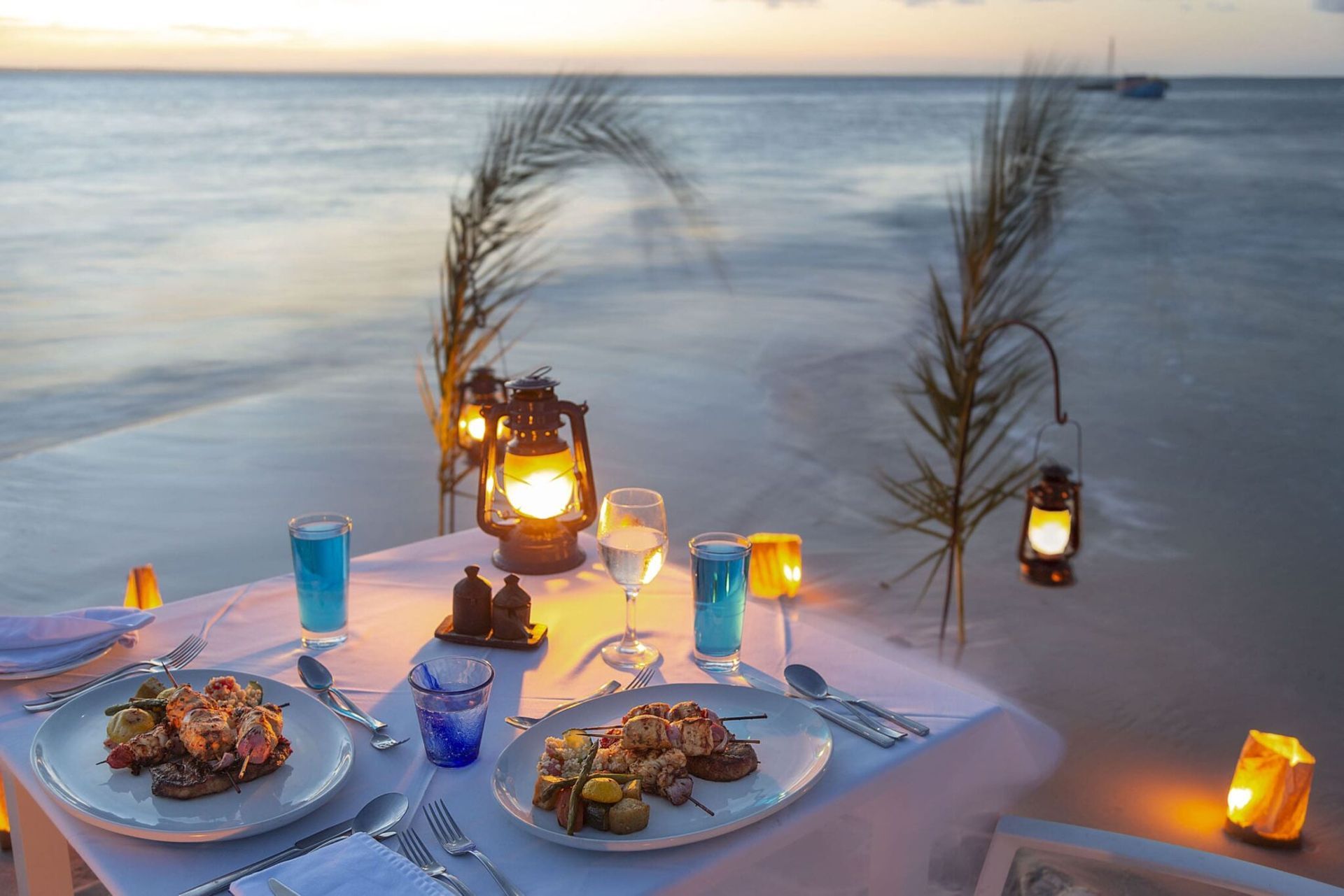 Romantic beach dinner table at sunset with lanterns, plates of food, and ocean view