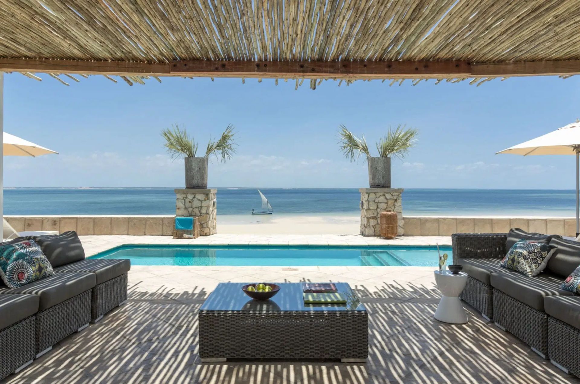 Oceanfront patio with wicker seating, shaded pergola, pool, and sailboats on a calm blue sea.