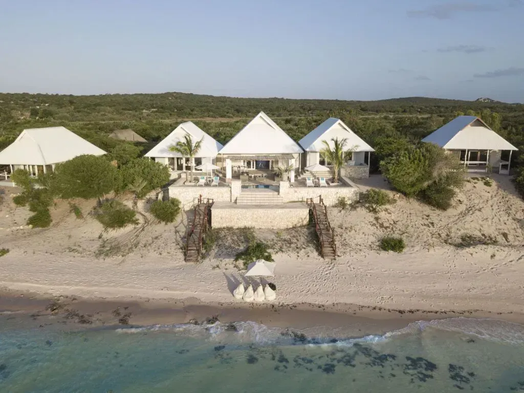 Aerial view of white beach villas on a sandy shoreline beside calm blue water and green vegetation