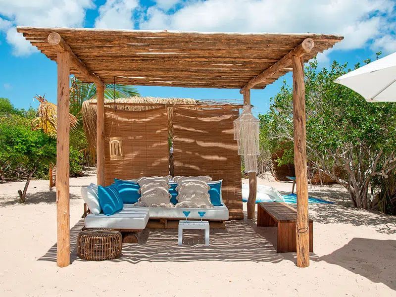 Wooden cabana with blue cushions and a white table on a sandy beach under a clear sky