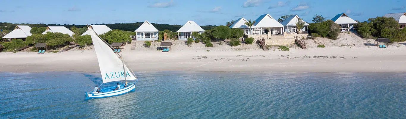 Blue sailboat on calm water near a sandy beach with white-roofed huts and palm trees