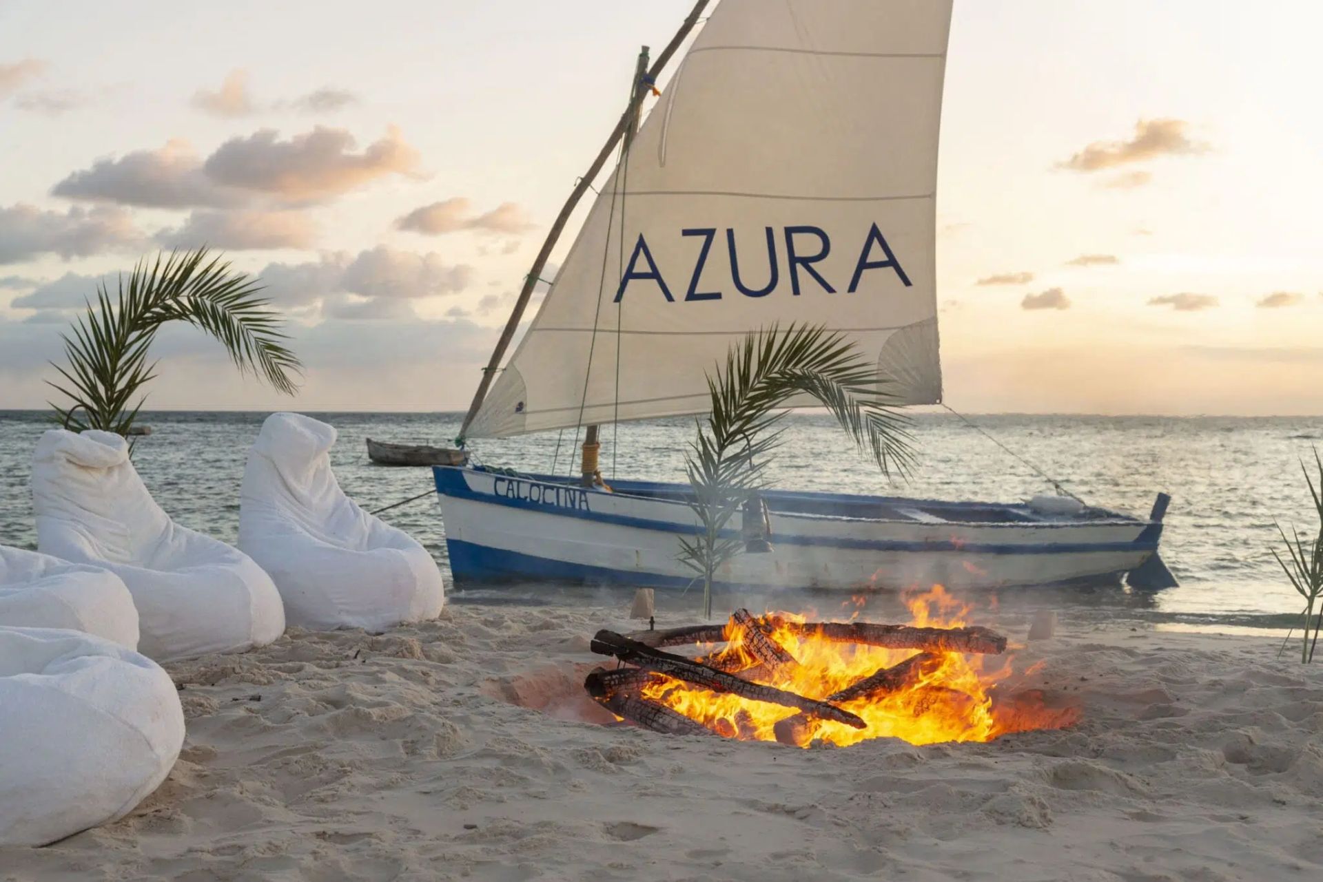 Sailboat with AZURA sail beside a beach bonfire at sunset, with white lounge chairs and palm fronds