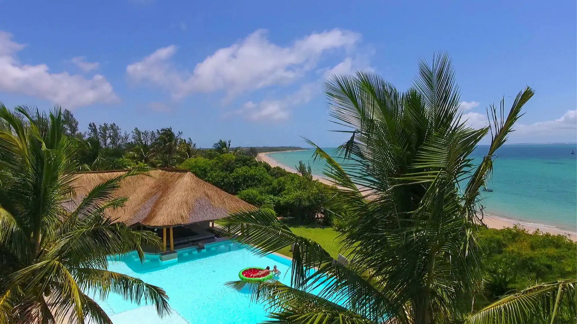 Tropical resort pool with thatched-roof huts, palm trees, and a turquoise ocean under a blue sky