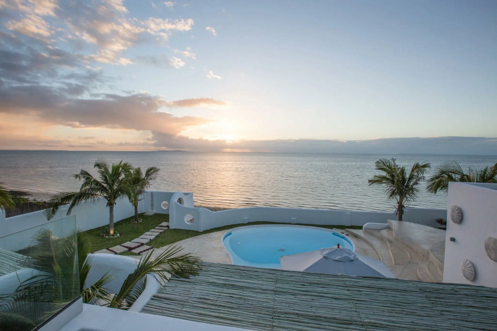 Oceanfront resort at sunset with a pool, palm trees, and calm water reflecting the sky.