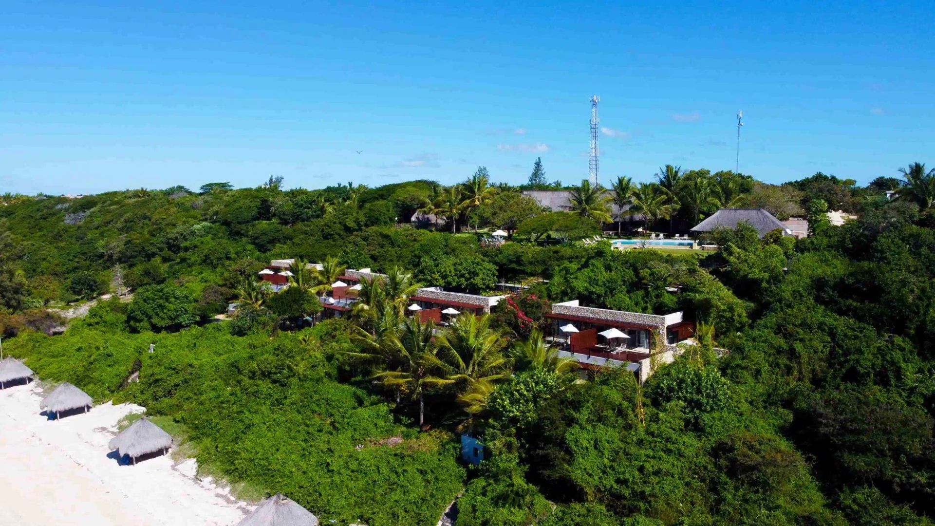 Tropical resort buildings surrounded by lush green trees beside a white sandy beach