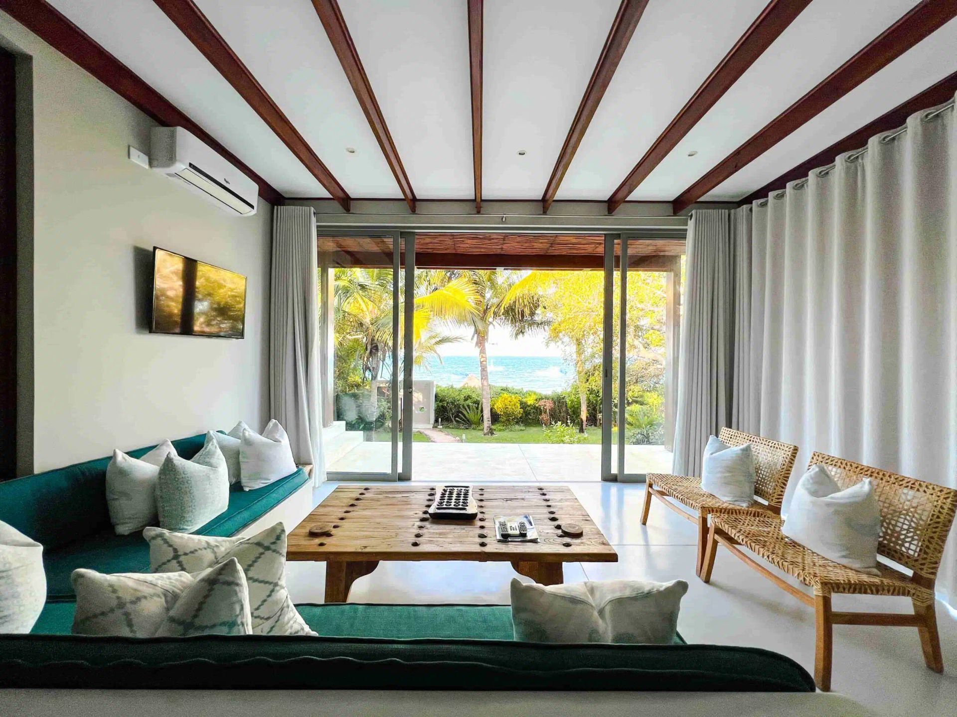 Bright modern living room with teal sofa, wooden table, wicker chairs, and open glass doors to a sunny garden