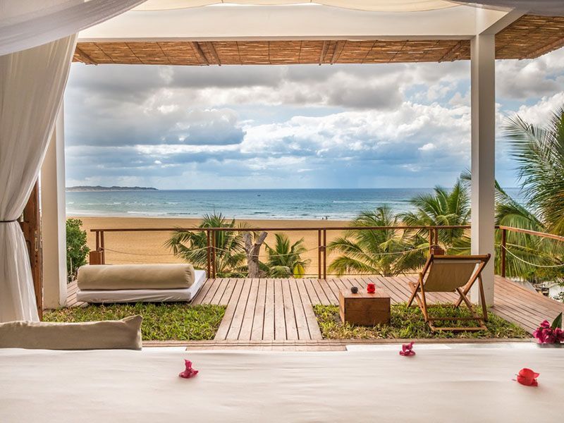 Open-air beach lounge with ocean view, white curtains, tan seating, and pink flowers on a patio