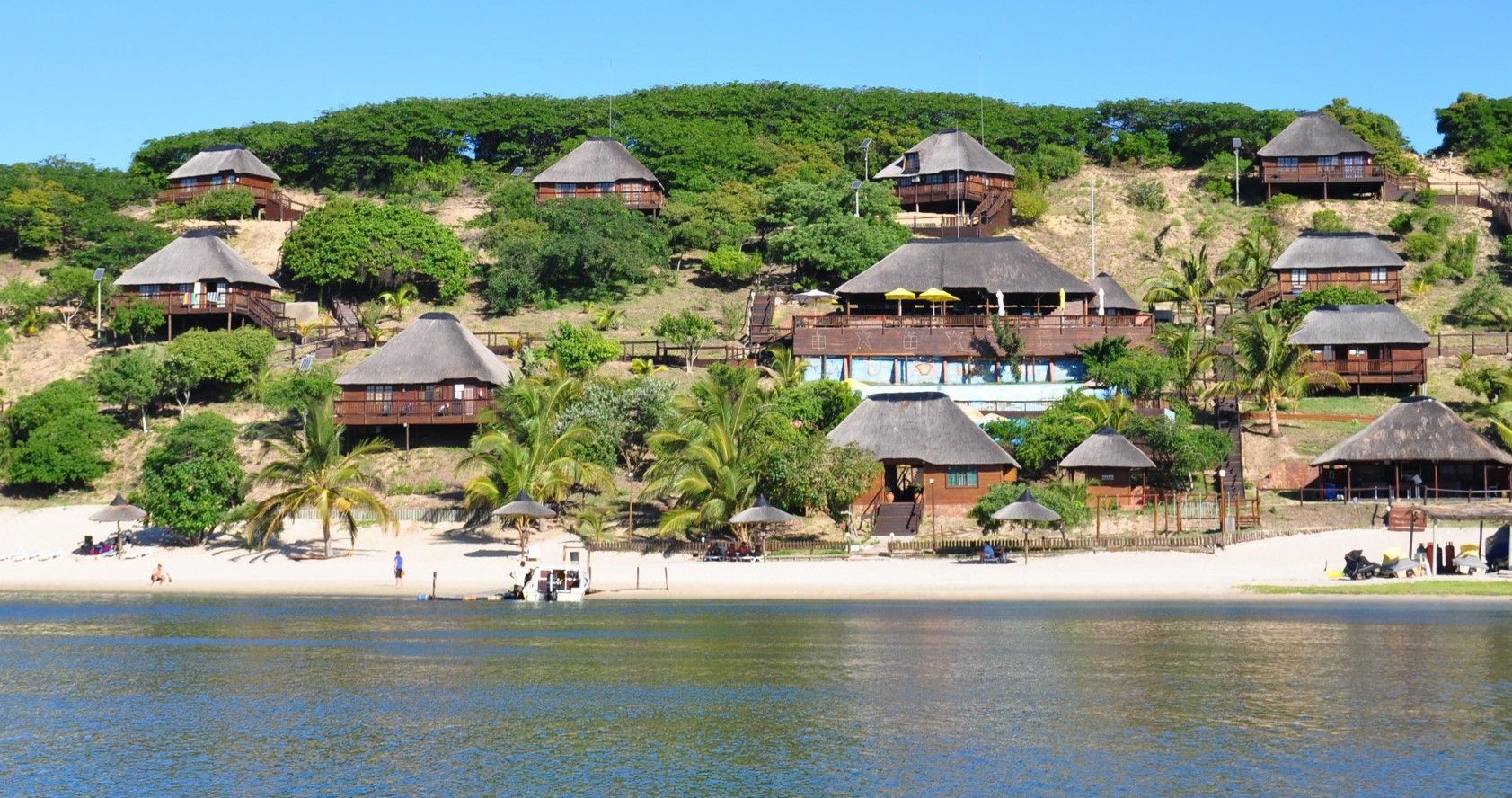 Tropical lakeside resort with thatched-roof huts on a green hillside above a sandy beach and calm water