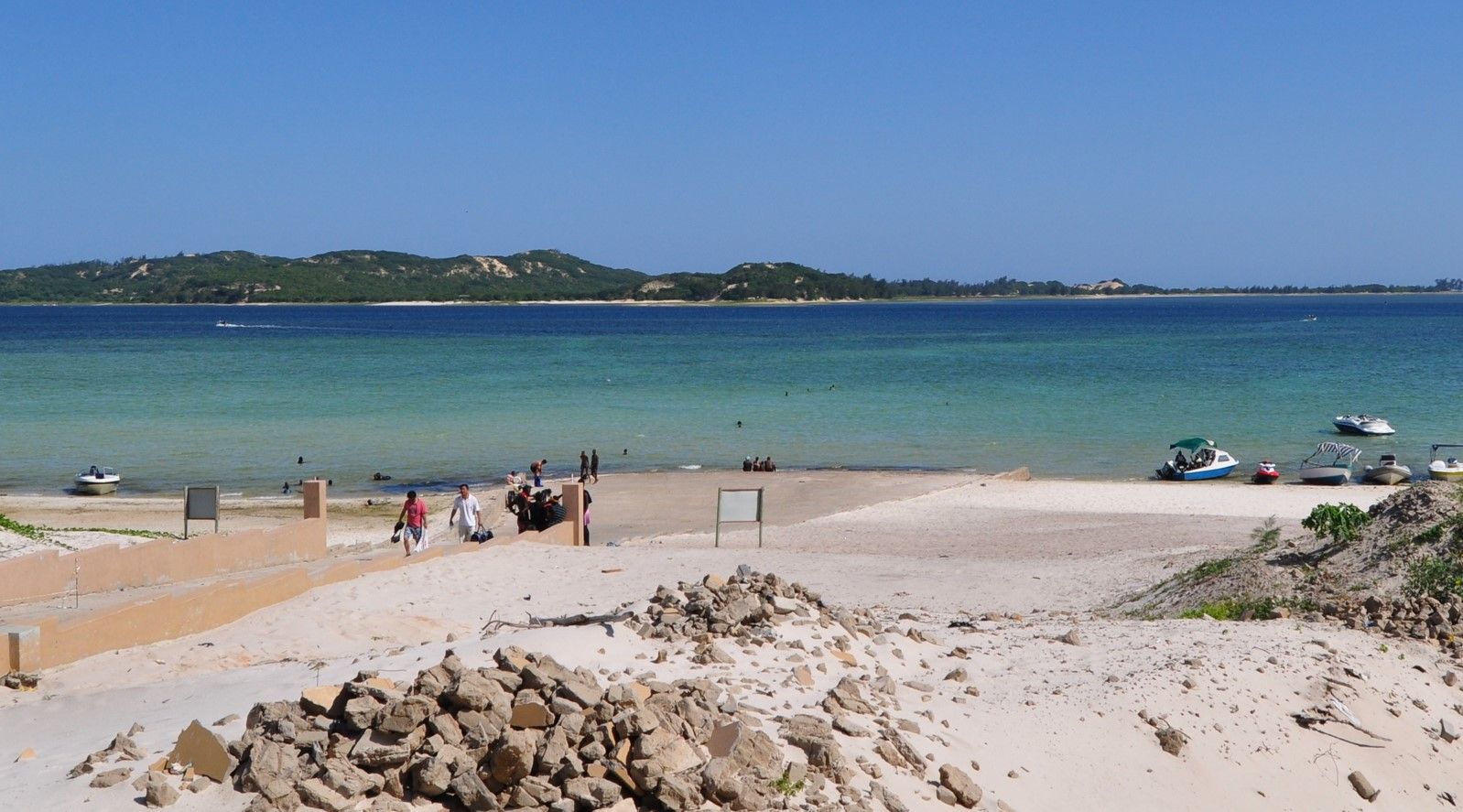 Sunny beach with white sand, turquoise water, and a few people near the shoreline, with a small island in the distance