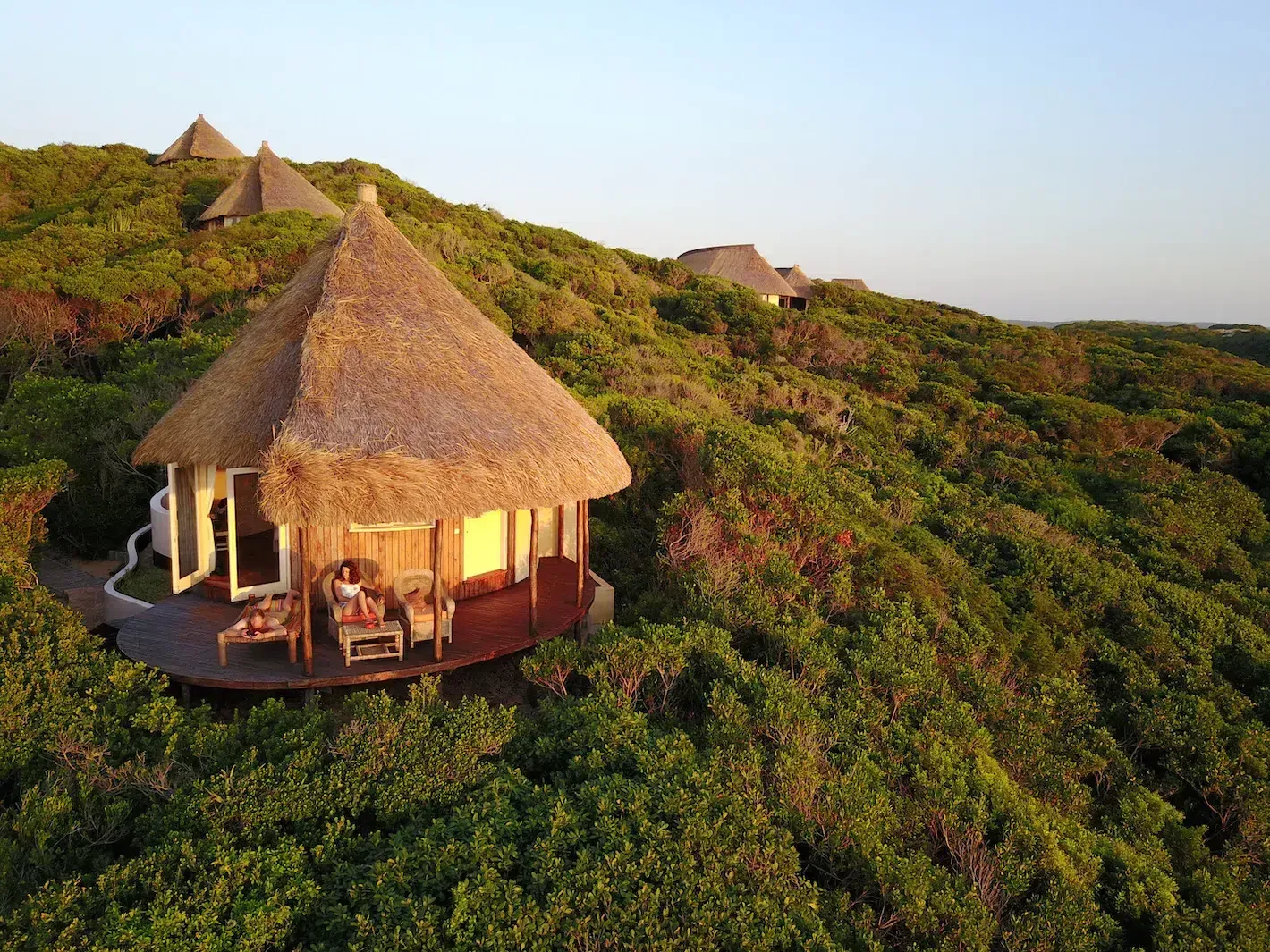 Thatched eco-lodge on a lush green hillside at sunset, with several huts in the distance.