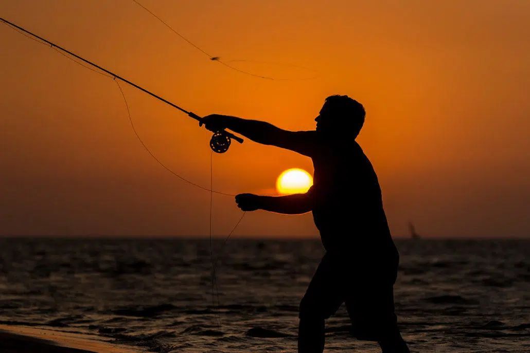 Silhouette of a person fishing at sunset by the ocean, with a glowing orange sky.