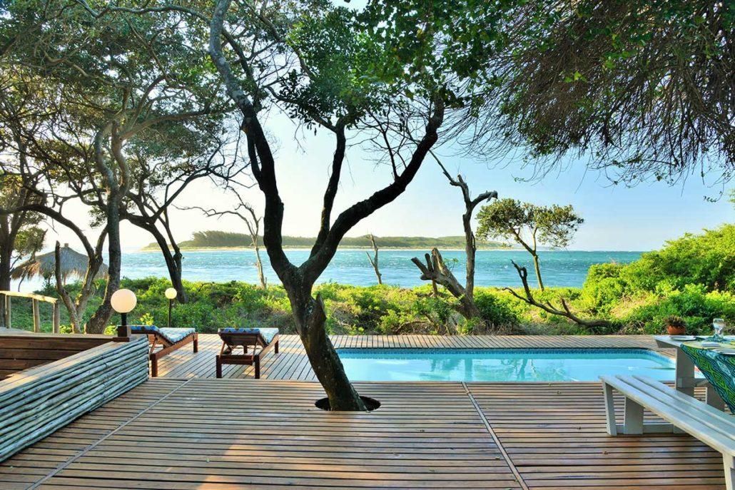 Oceanfront deck with pool, trees, and lounge seating overlooking the beach.