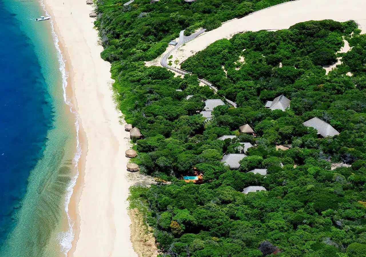 Aerial view of a tropical beach with turquoise water, white sand, and dense green forest with thatched-roof huts.