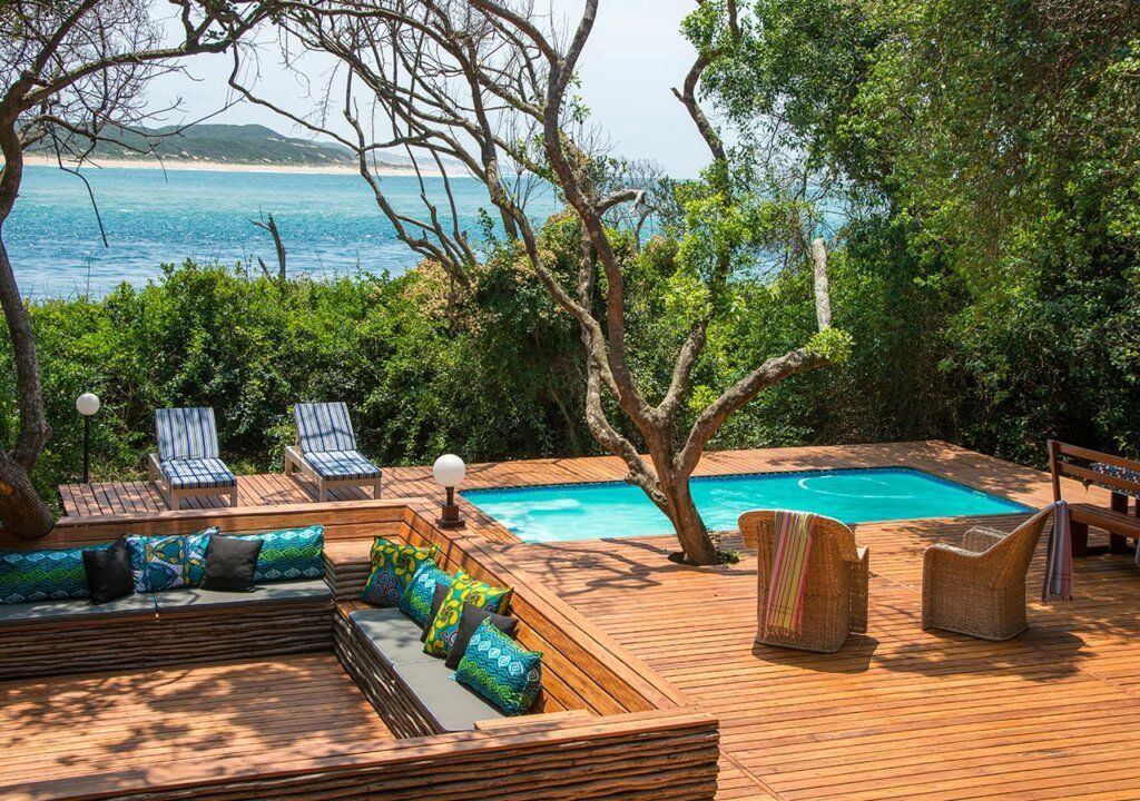Oceanfront patio with pool, lounge chairs, and wooden deck surrounded by lush greenery