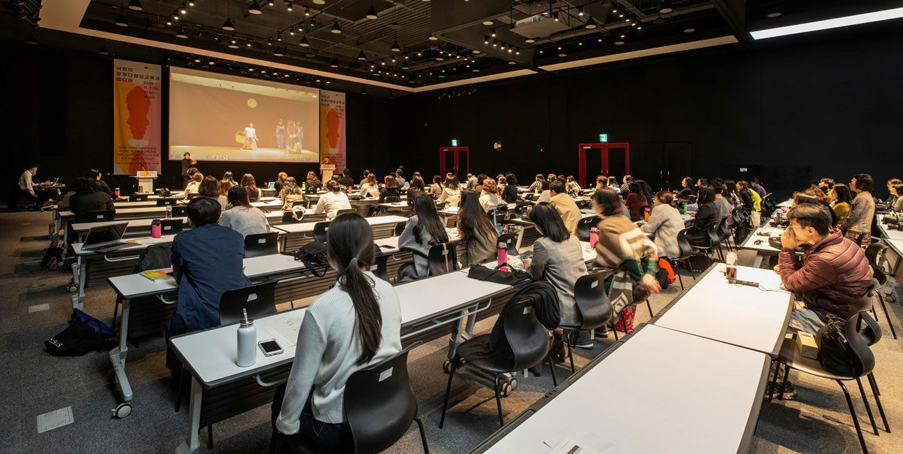 Large conference hall with audience seated at long tables facing a projected presentation screen and stage.