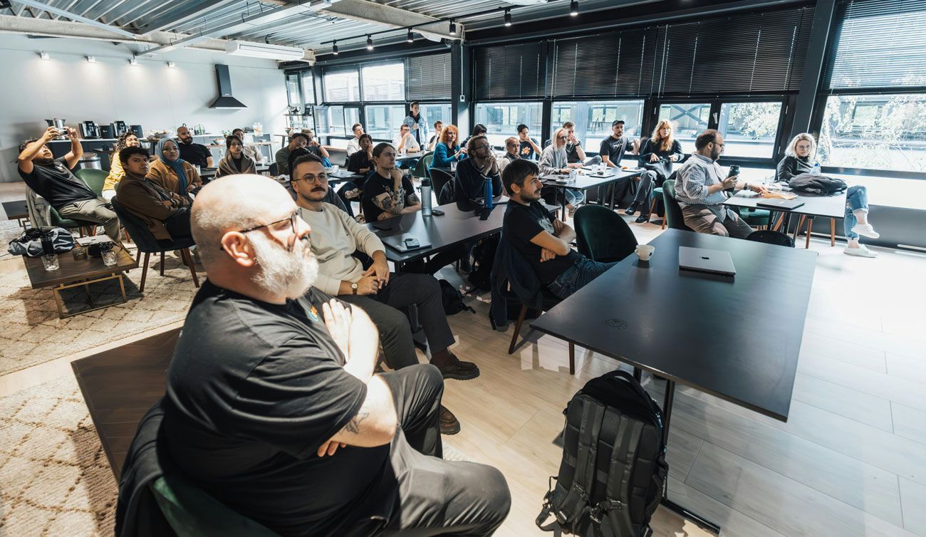 Audience seated in a bright conference room, listening to a presentation at a large screen