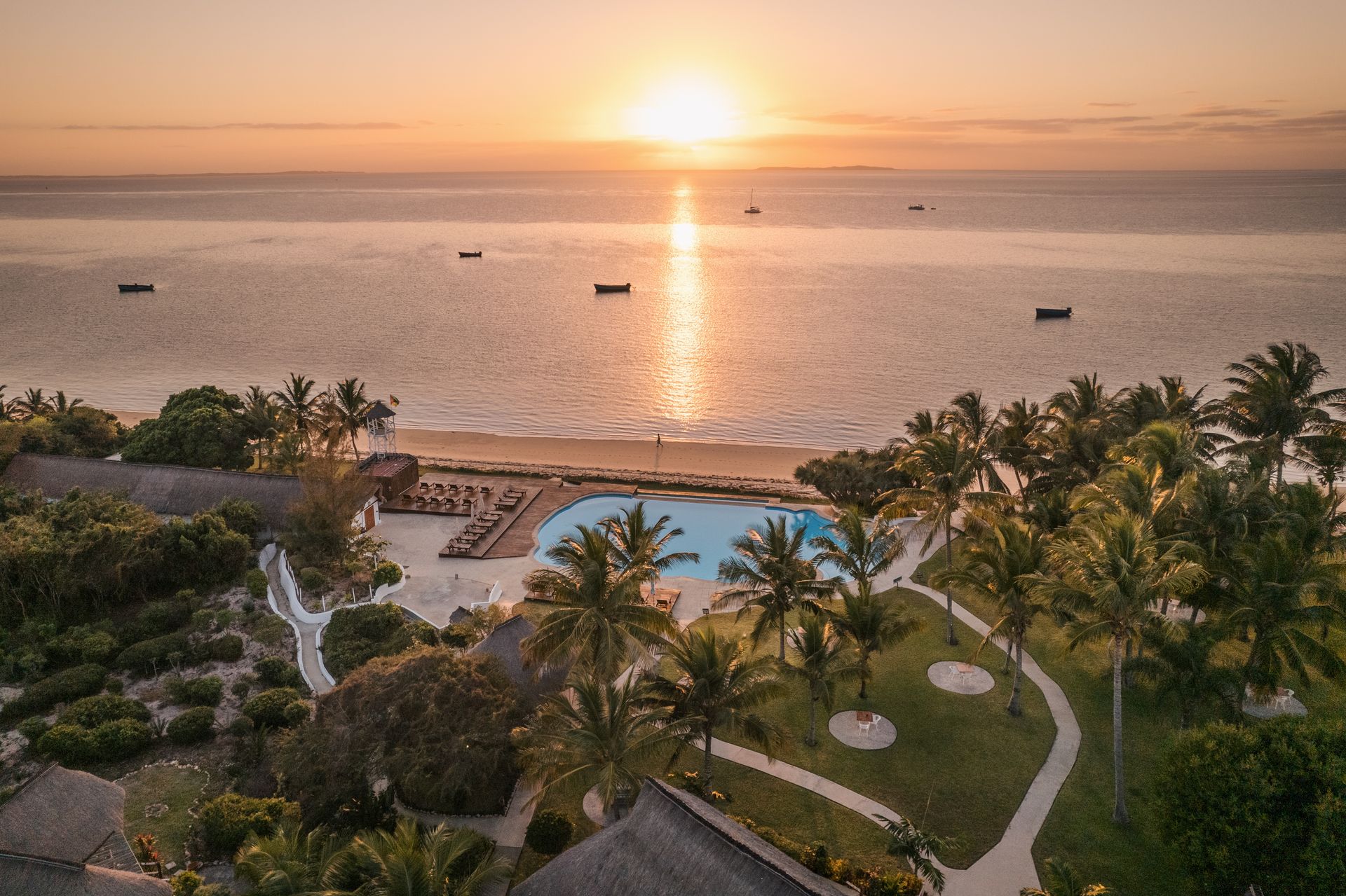 Sunset over a tropical beach resort with palm trees, pool, and boats on calm water