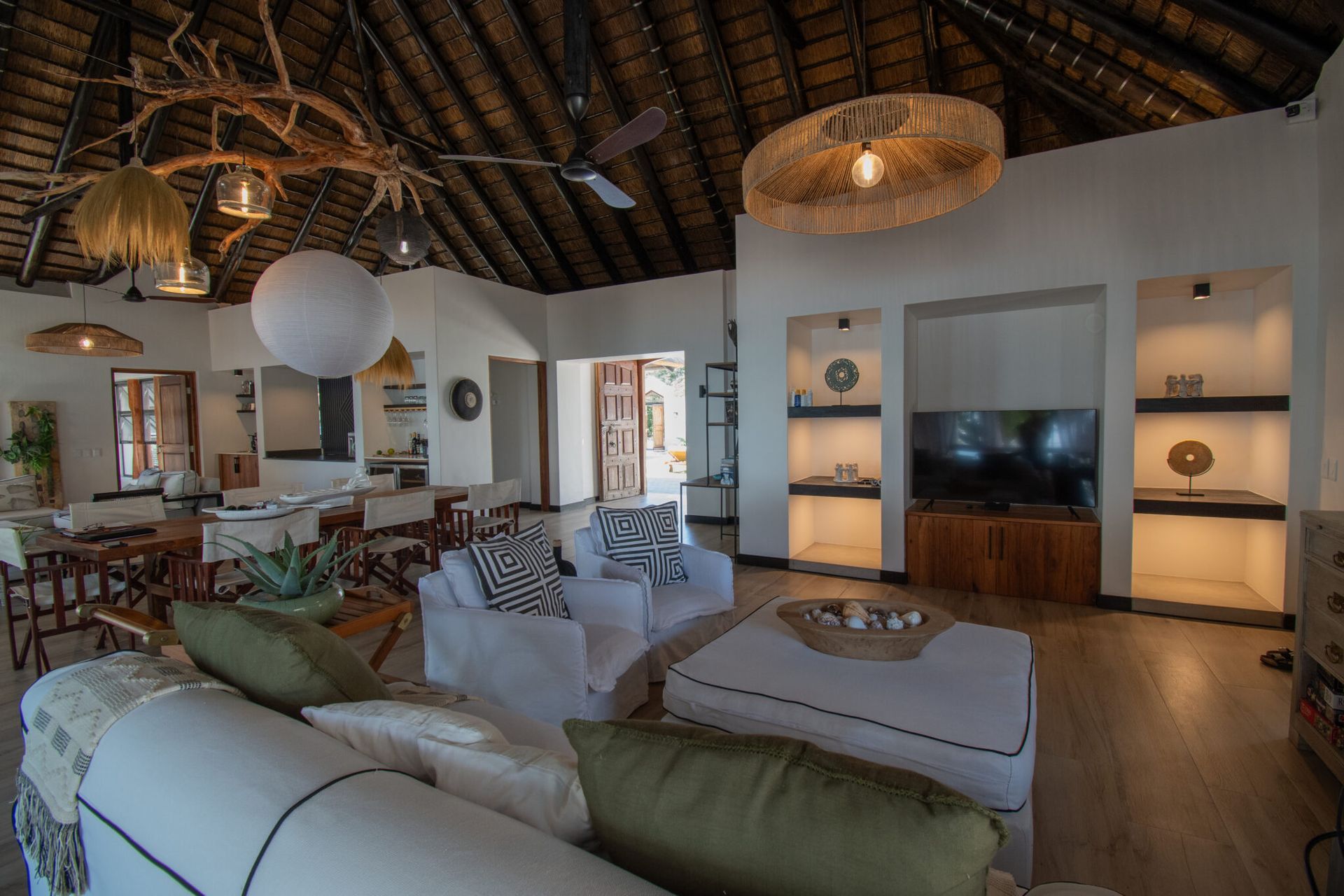 Open-air living room with thatched ceiling, white sofas, wooden furniture, and a TV on a wall unit.
