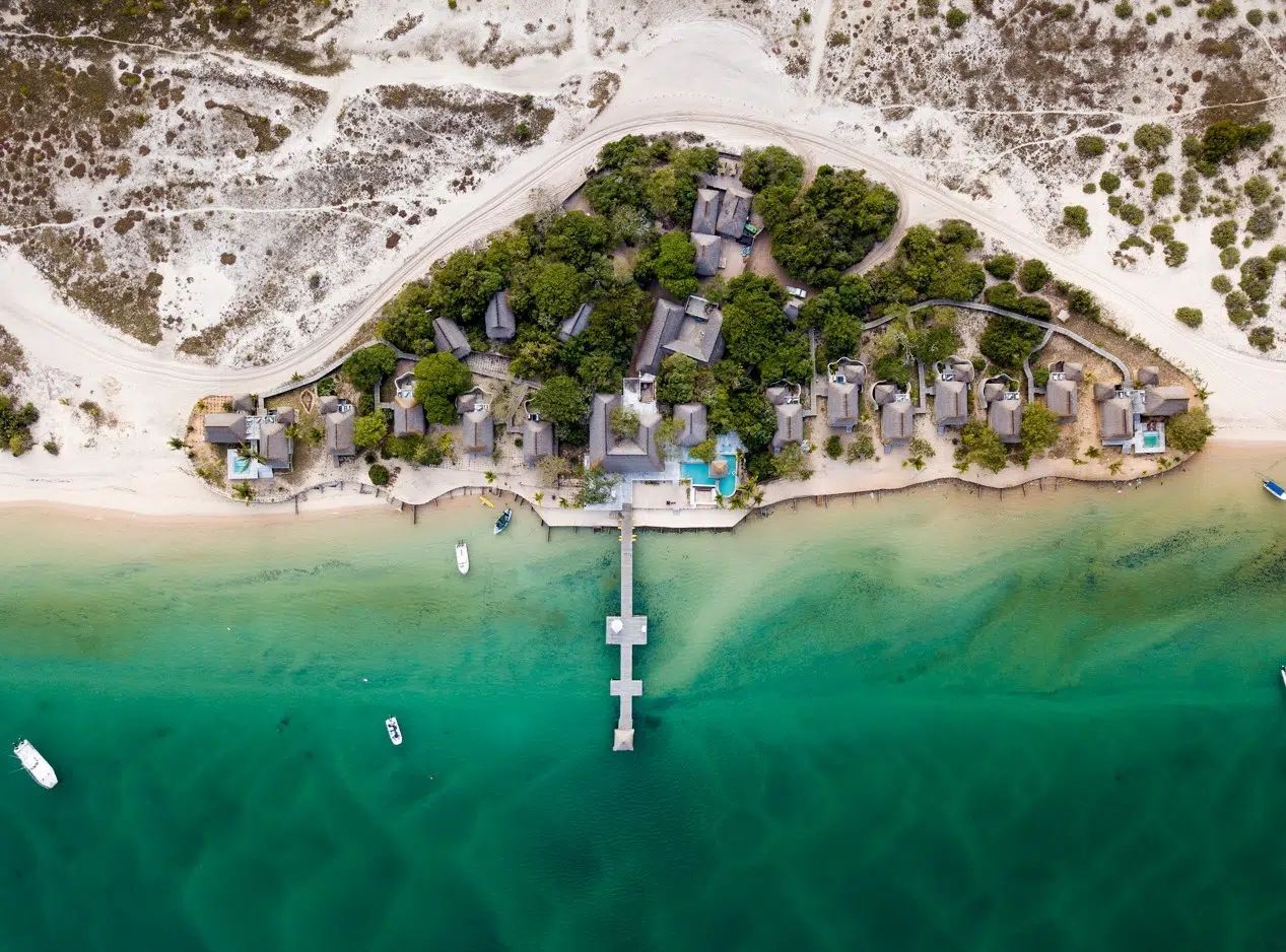 Aerial view of a tropical beach with turquoise water, a pier, and palm-covered buildings onshore