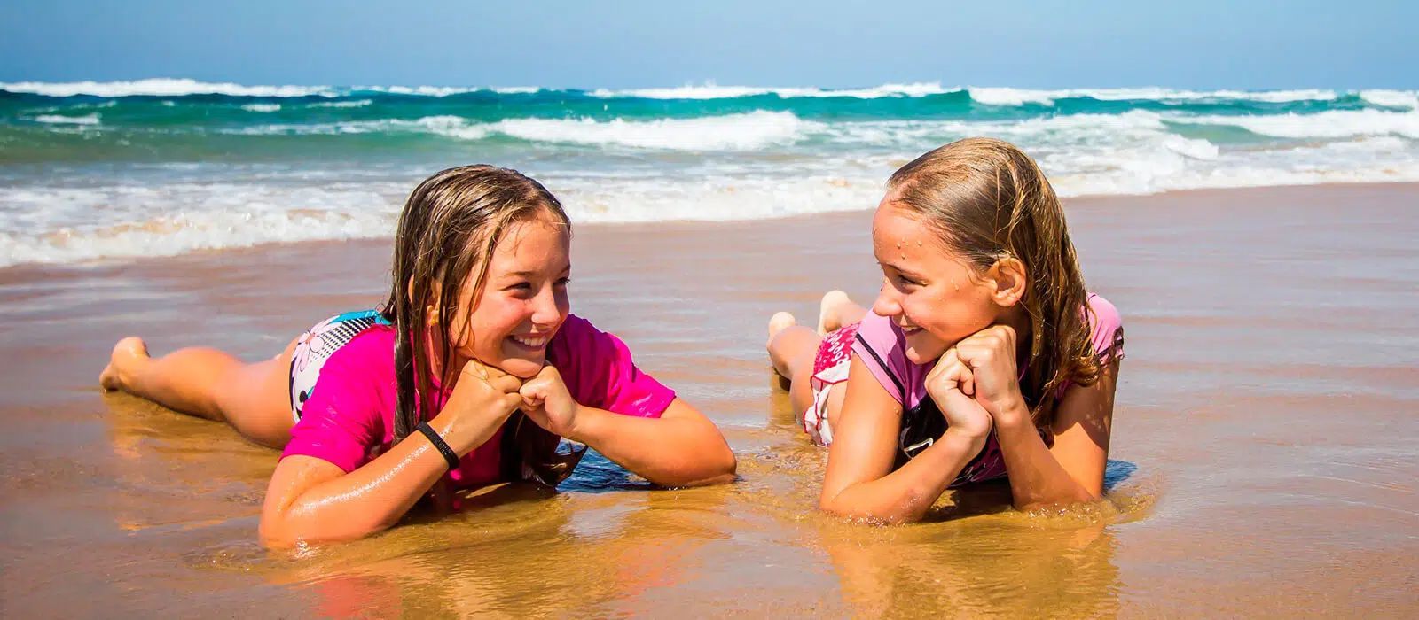 Two children lying in shallow surf, smiling and talking on a sunny beach