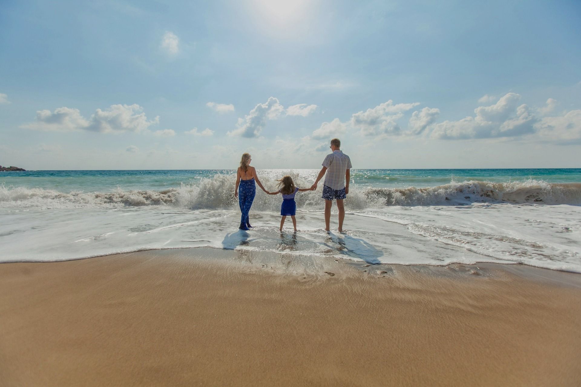 Three people hold hands walking into ocean waves on a sunny beach