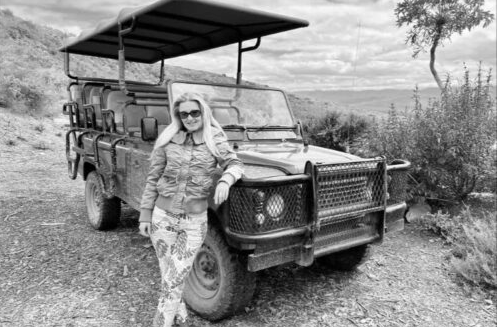Person in sunglasses leaning on a safari vehicle on a dirt road in a rural landscape.