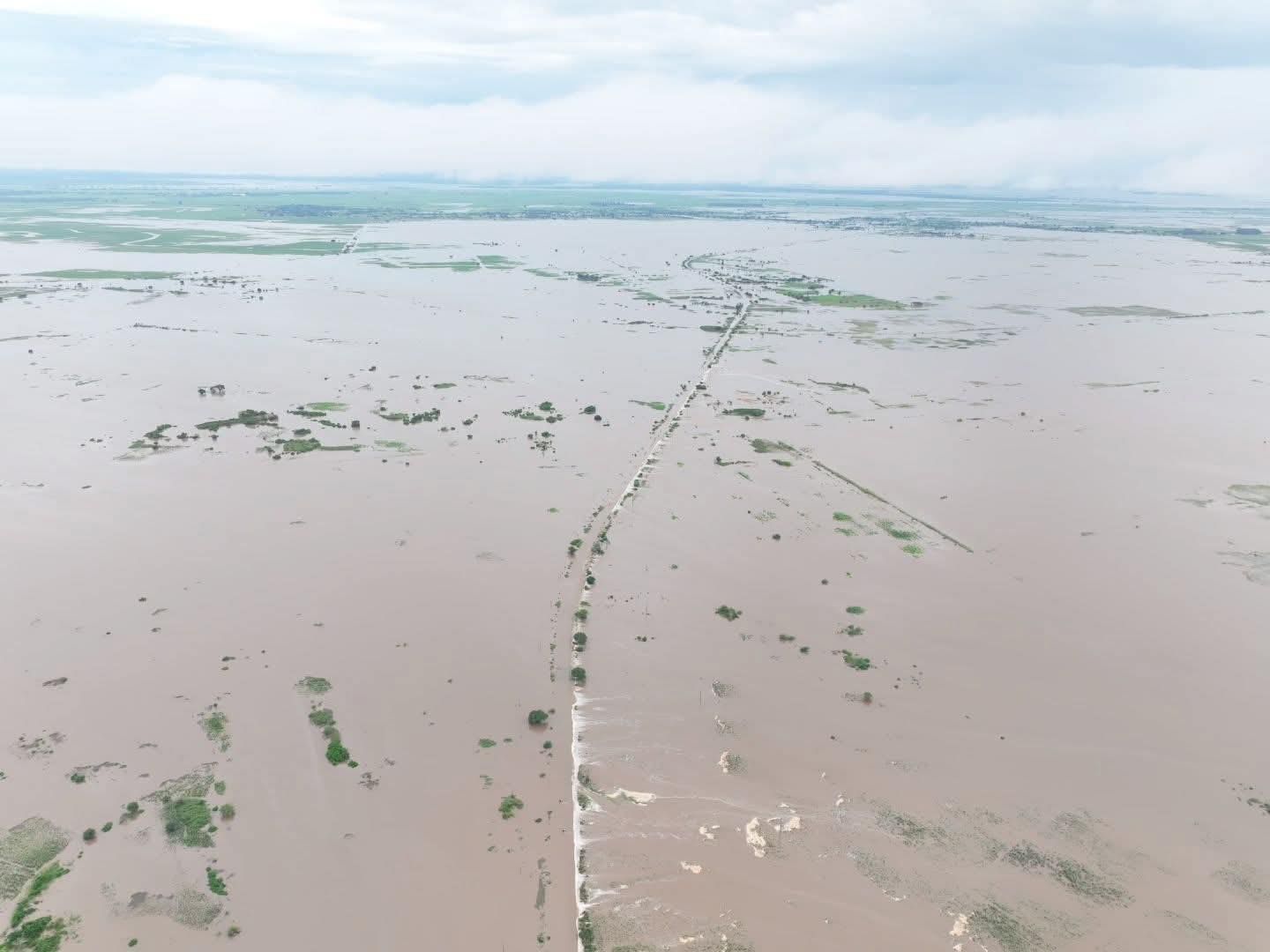A flooded landscape with muddy water, scattered vegetation, and a narrow road stretching into the distance.