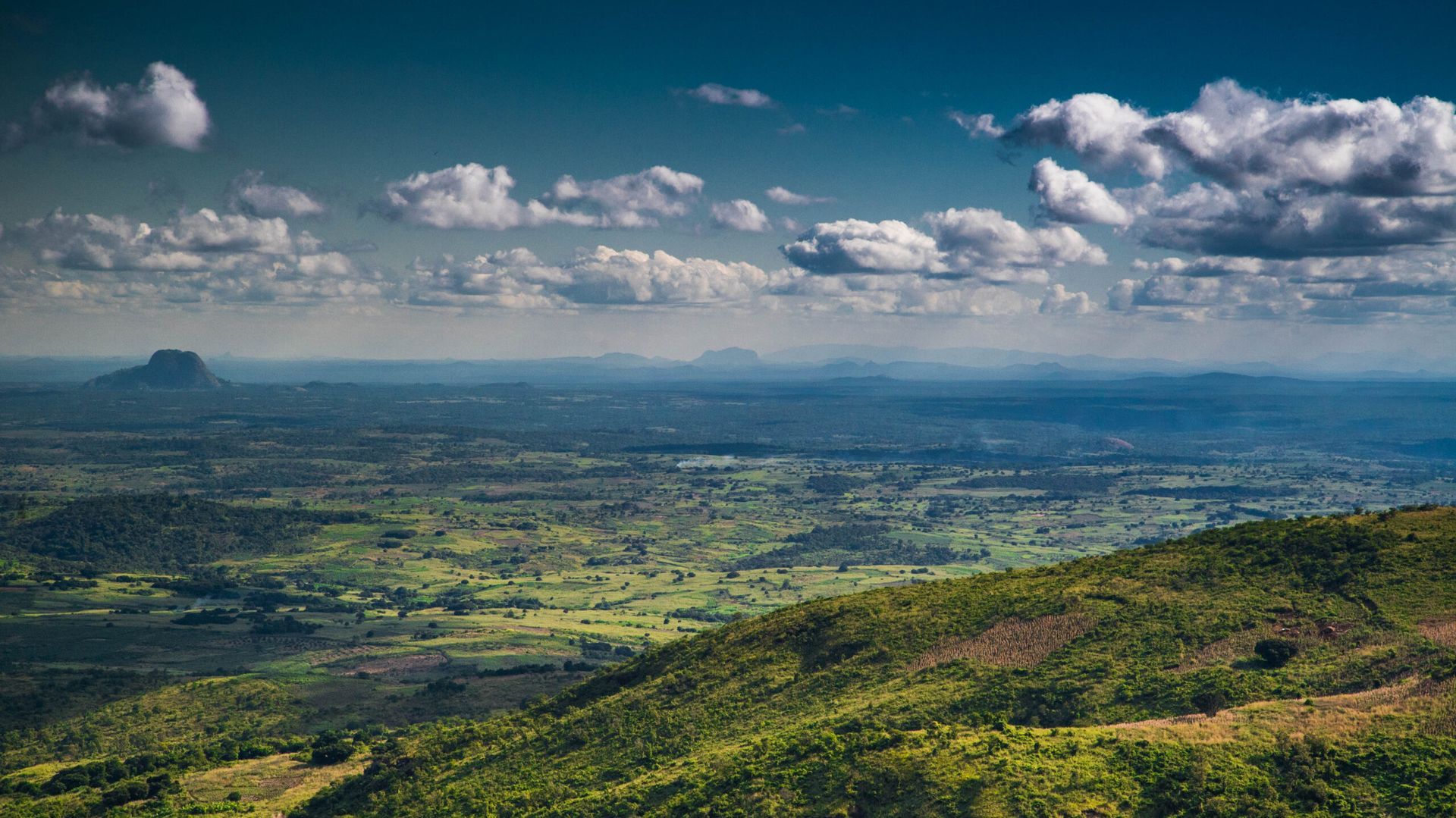 Wide green valley under a cloudy blue sky, viewed from a hillside with distant mountains.
