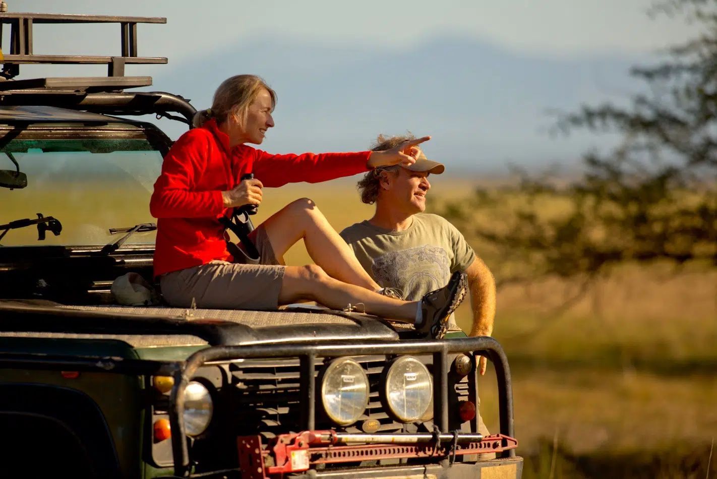 Two people sitting on a safari vehicle roof in a grassy field, one holding binoculars