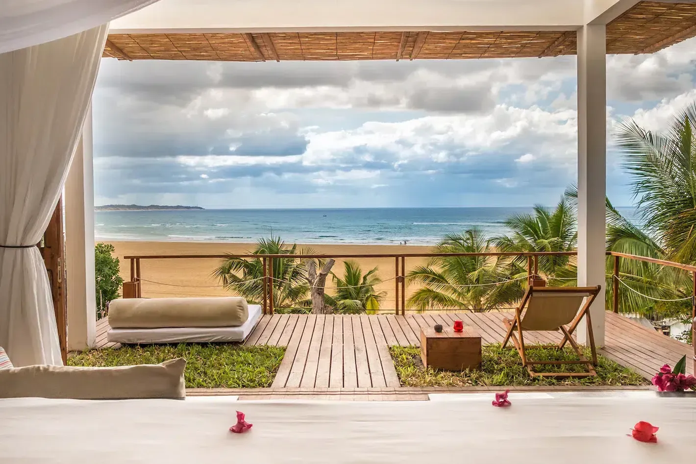 Beachfront patio with lounge chairs, white curtains, and ocean view under a cloudy sky