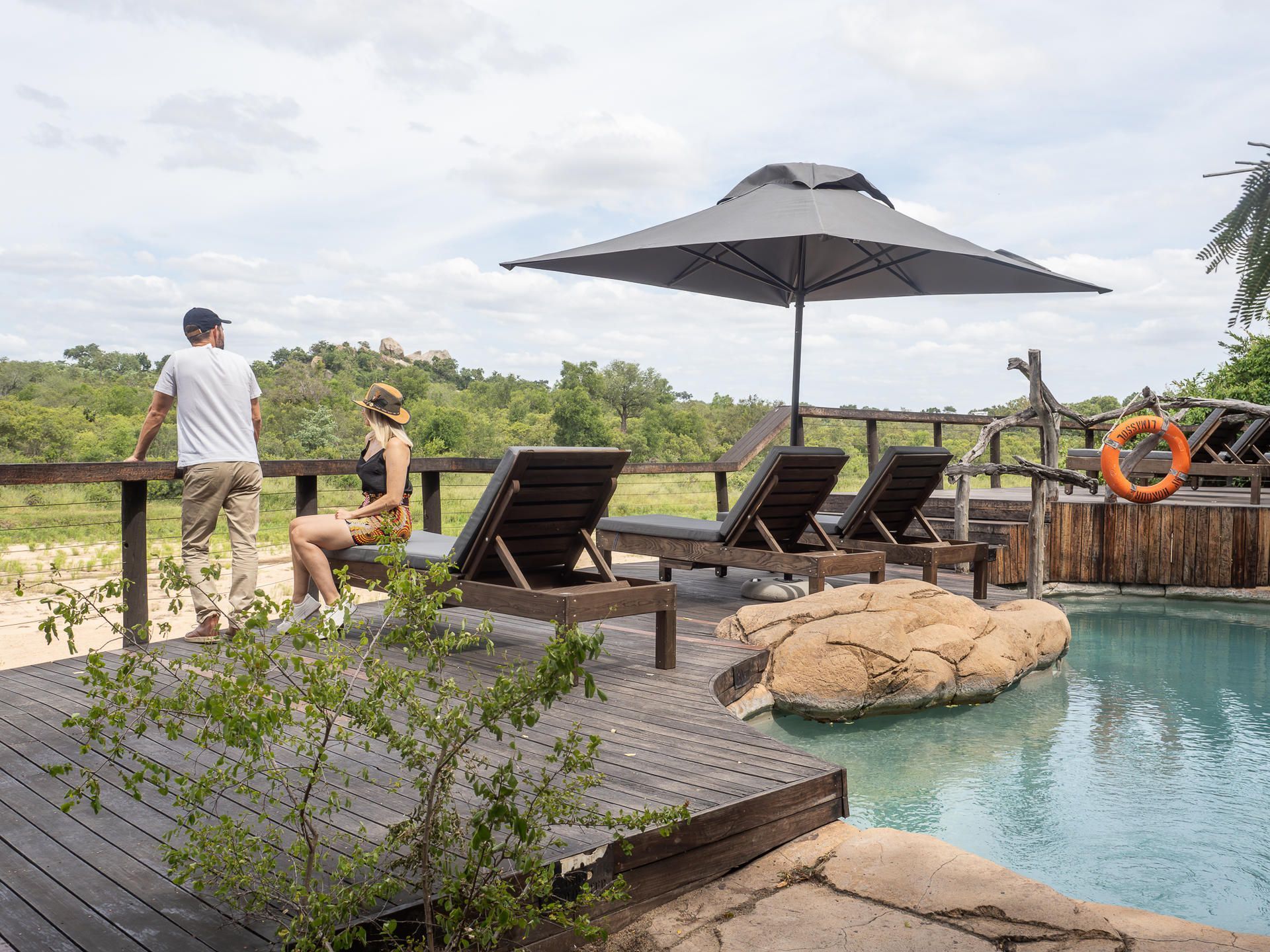 Resort poolside deck with lounge chairs, umbrellas, and a person standing by a railing overlooking greenery