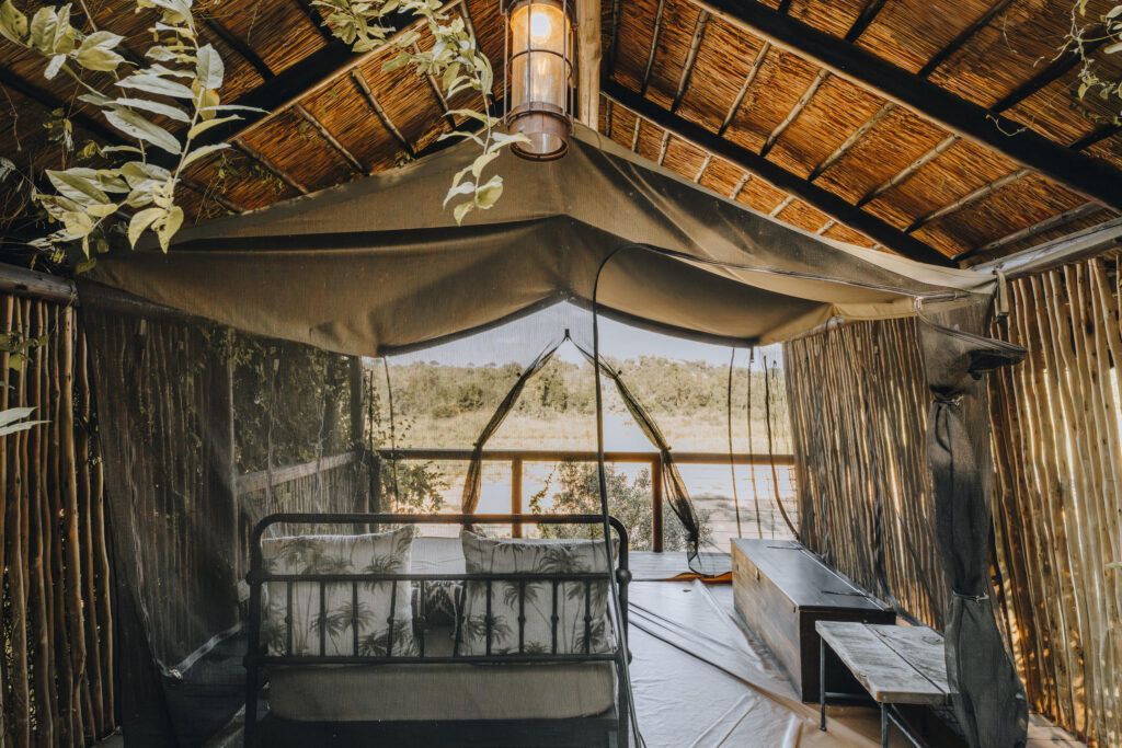 Rustic thatched hut interior with a bed, woven walls, and a view of the outdoors beyond open curtains