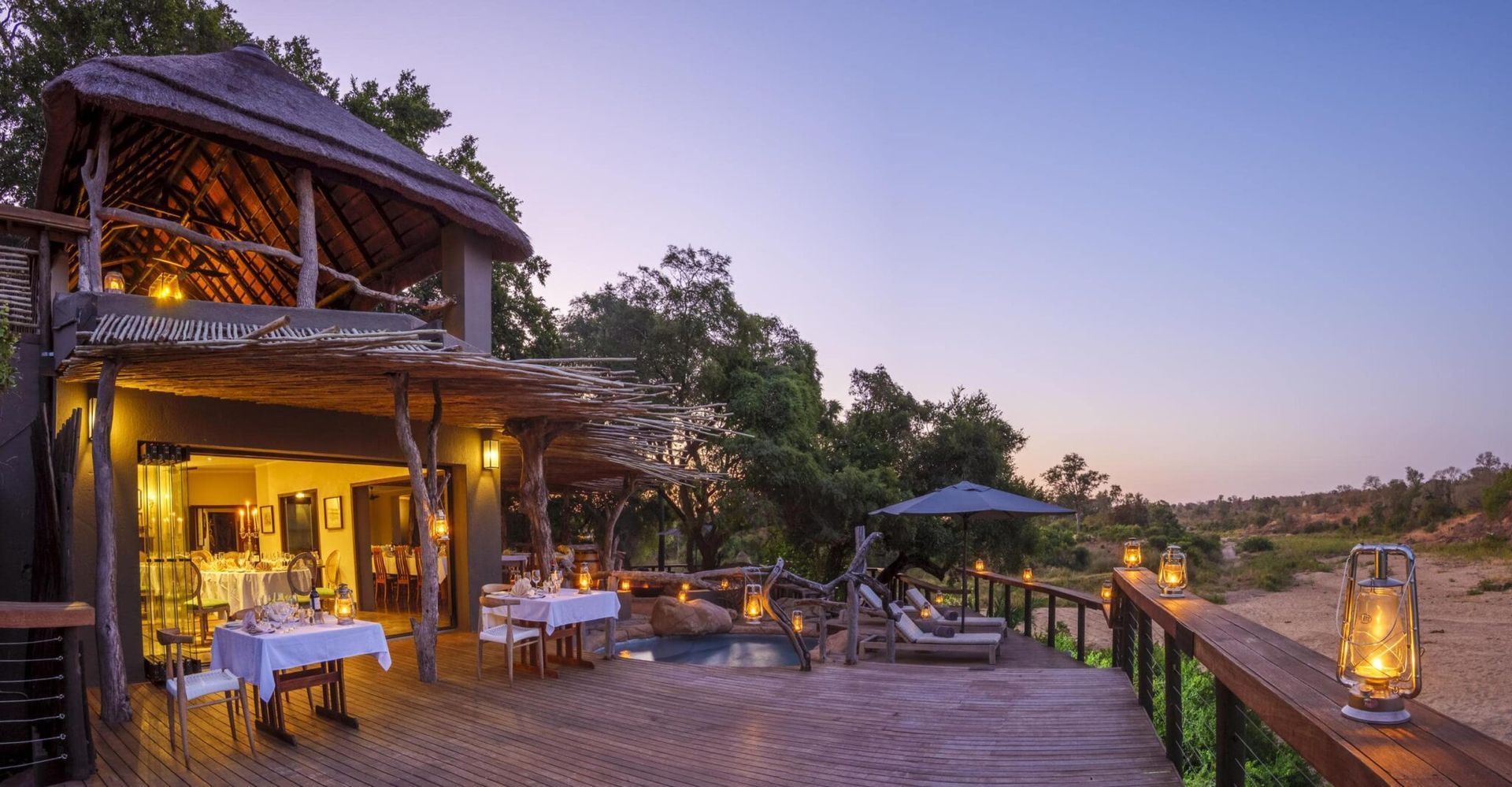 Rustic riverside lodge at dusk with warm lights, outdoor dining tables, and a wooden deck overlooking the water