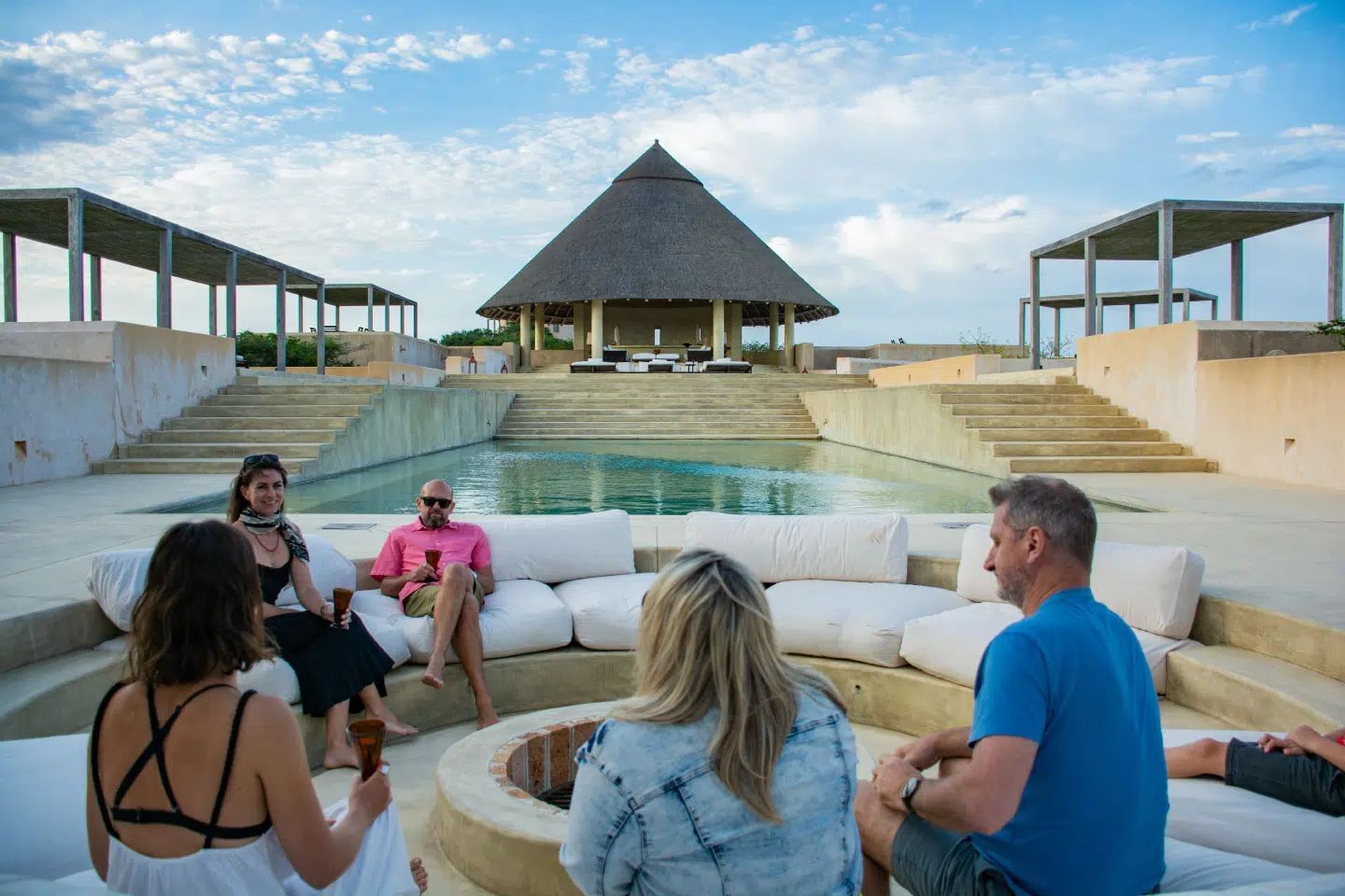 Four people sit on a circular lounge around a fire pit by a resort pool, with a large thatched-roof building in the back.