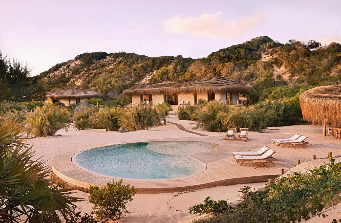 A sandy resort area with a pool surrounded by low shrubs, lounge chairs, and thatched-roof villas against a hillside.